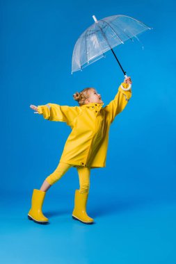 Mavi arka planda şeffaf bir şemsiyeyle duran yağmurluk ve lastik çizmeli neşeli kız. Reklam metniniz için boşluk resmini yerleştirerek kopyalayın