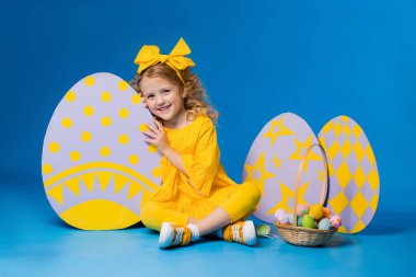 Sarı elbiseli küçük sarışın kız mavi arka planda büyük dekoratif paskalya yumurtalarıyla ayakta duruyor.