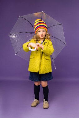 Sarı yağmurluk ve mor arka planda şeffaf şemsiye tutan renkli şapkalı hoş bir kız. Metnin ya da ilanın için boşluk resmini yerleştirerek kopyala