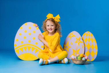Sarı elbiseli küçük sarışın kız mavi arka planda büyük dekoratif paskalya yumurtalarıyla ayakta duruyor.