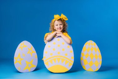 Mavi arka planda büyük dekoratif paskalya yumurtaları olan sarı elbiseli mutlu kız. Metnin ya da ilanın için boşluk resmini yerleştirerek kopyala