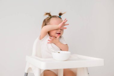 Beyaz tişörtlü şirin bir kız çocuğu yüksek sandalyede oturup lapa yiyor.