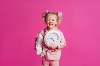 Açık pembe pijamalarla poz veren güzel bir kızın stüdyo fotoğrafı. Yastık, tüy ve saatle pembe arka planda duran neşeli bir kız bebek. Çocuk duyguları konsepti