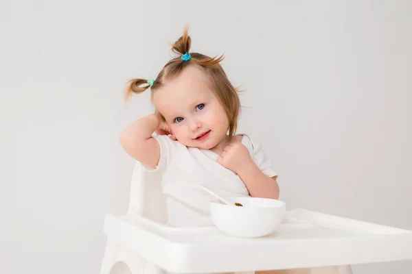 Cute baby girl in white t-shirt sitting in a high chair and eating porridge