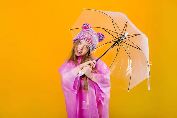 Photo of a little girl in a pink raincoat and  knitted hat holding an umbrella isolated on yellow background with a space for text