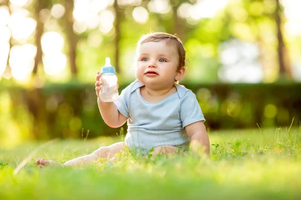 夏に草の上に座ってボトルから水を飲む青いボディスーツのかわいい幼児 — ストック写真