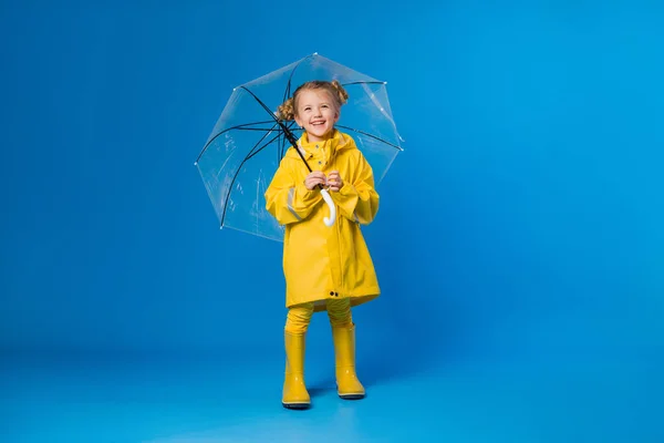 Cheerful girl in a raincoat and rubber boots standing on a blue background with a transparent umbrella. Copy space picture with placement for your text of advertisement