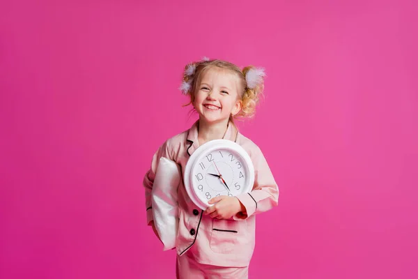 Açık pembe pijamalarla poz veren güzel bir kızın stüdyo fotoğrafı. Yastık, tüy ve saatle pembe arka planda duran neşeli bir kız bebek. Çocuk duyguları konsepti