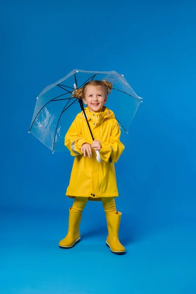 Cheerful girl in a raincoat and rubber boots standing on a blue background with a transparent umbrella. Copy space picture with placement for your text of advertisement