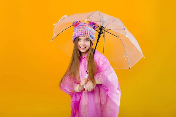 Photo of a little girl in a pink raincoat and  knitted hat holding an umbrella isolated on yellow background with a space for text