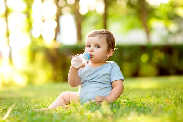 夏に草の上に座ってボトルから水を飲む青いボディスーツのかわいい幼児 — ストック写真