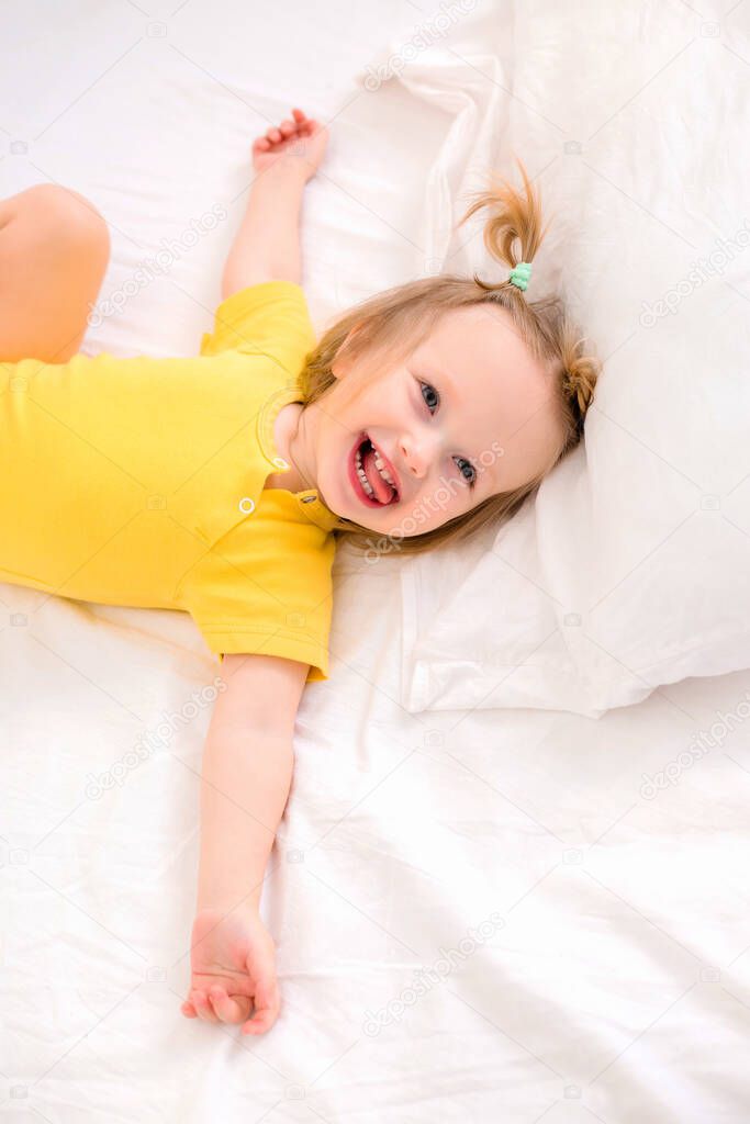 Toddler girl in yellow bodysuit smiles, lies on a white bed  
