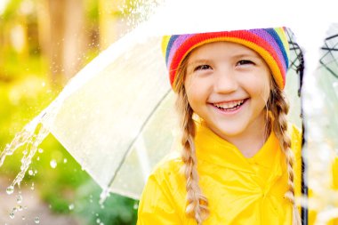 Sarı yağmurluk ve şeffaf şemsiyeli mutlu kız yağmurda eğleniyor.