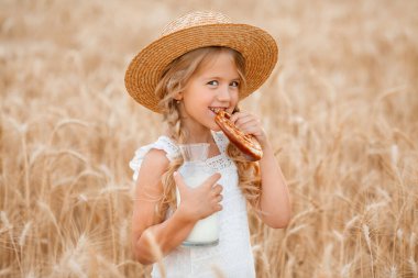 Hasır şapkalı ve beyaz elbiseli küçük sarışın kız ekmek yiyor ve buğday tarlasında süt içiyor. Çevre dostu tarım ürünleri kavramı