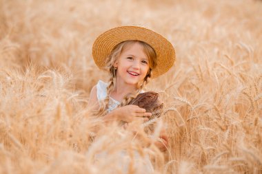Hasır şapkalı ve beyaz elbiseli küçük sarışın bir kız buğday tarlasındayken bir somun taze ekmek tutuyor. Doğal tarım ürünleri ve eko yaşam konsepti