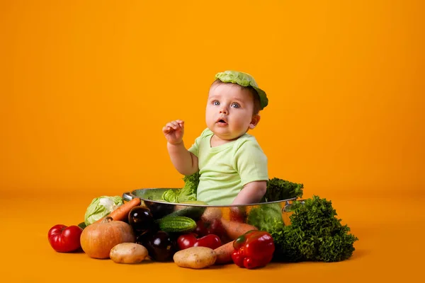 赤ちゃんの男の子は キャベツの葉を帽子として新鮮な野菜を入れた大きなボウルに座っています オレンジの背景に孤立した画像 — ストック写真