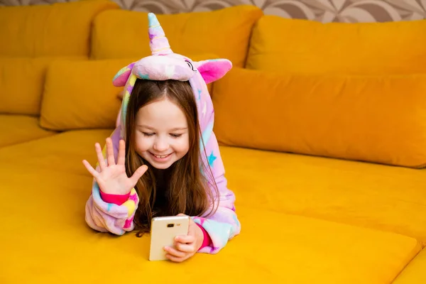 Little girl in unicorn onesie lies at home on a couch with a smartphone. Isolated on orange background, video call or mobile game playing concept