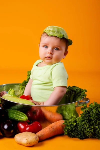 赤ちゃんの男の子は キャベツの葉を帽子として新鮮な野菜を入れた大きなボウルに座っています オレンジの背景に孤立した画像 — ストック写真