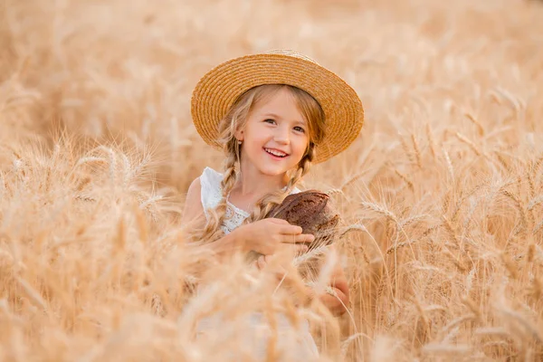 Hasır şapkalı ve beyaz elbiseli küçük sarışın bir kız buğday tarlasındayken bir somun taze ekmek tutuyor. Doğal tarım ürünleri ve eko yaşam konsepti