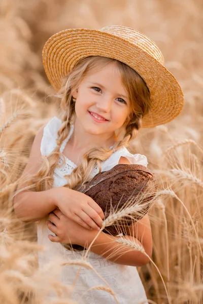 Hasır şapkalı ve beyaz elbiseli küçük sarışın bir kız buğday tarlasındayken bir somun taze ekmek tutuyor. Doğal tarım ürünleri ve eko yaşam konsepti
