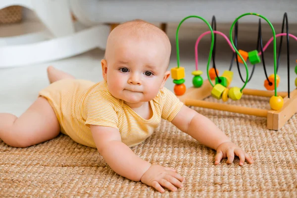 黄色のストライプのボディスーツのかわいい幼児は カーペットの上で発達したおもちゃで遊んで座っています 子供の発達の概念 — ストック写真