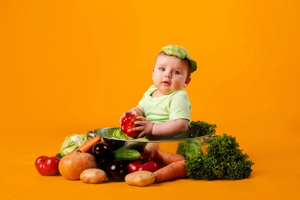 赤ちゃんの男の子は キャベツの葉を帽子として新鮮な野菜を入れた大きなボウルに座っています オレンジの背景に孤立した画像 — ストック写真