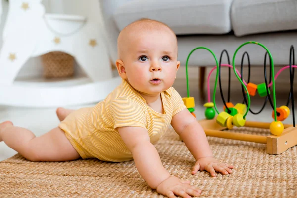 黄色のストライプのボディスーツのかわいい幼児は カーペットの上で発達したおもちゃで遊んで座っています 子供の発達の概念 — ストック写真