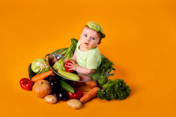 緑色のボディスーツの赤ん坊は黄色の背景に野菜と金属製のボウルに座っています 農産物の概念やテキストの空間 — ストック写真