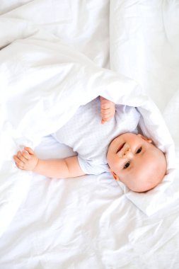 Beyaz battaniyeye sarılı tatlı erkek bebek yatakta uzanıyor ve mutlu bir şekilde gülümsüyor.