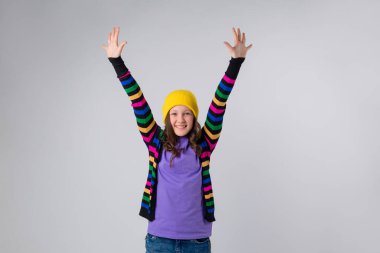 Sarı şapkalı genç kız, çok renkli süveter ve mor tişört. Açık gri arka planda izole edilmiş, gençlerin duygu konsepti..