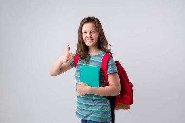 Sırt çantası ve elinde not defteriyle gülümseyen bir okul çocuğu. Açık gri arka planda izole edilmiş, gençlerin duygu konsepti..
