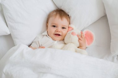 Sevimli mutlu küçük çocuk pelüş oyuncakla yatakta yatıyor.