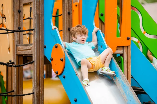 Cheerful redhead boy plays at colorful slide and climbing playground tower