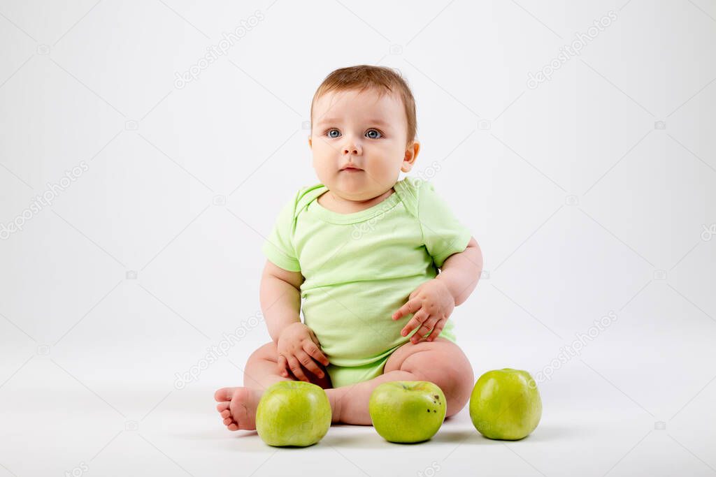 Cute toddler in green bodysuit sits  and plays with green apples. Isolated image on white background