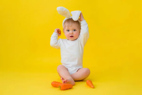 Baby in a white bodysuit with rabbit ears on his head eats carrots, sits on a yellow background with vegetables. baby in the form of an Easter bunny, space for text