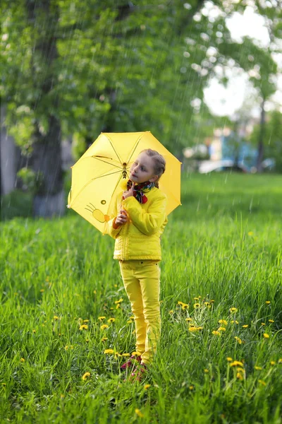 Funny cute girl wearing yellow coat holding colorful umbrella playing in the garden by rain and sun weather on a warm autumn or summer day