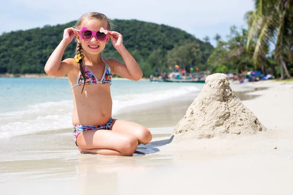Güneş gözlüğü ve denizde mayo giyen küçük mutlu bir kız. Rahatlama ve tatil kavramı. Deniz kıyısında kum oynayan kız