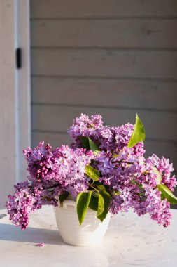 Beyaz bir vazodaki açık mor leylak demeti, yeşil bir bahçedeki bir masanın üzerine yerleştirilmiş. Taze ve huzurlu bir sahne Bir bahar gününün romantik güzelliğini yakalıyor