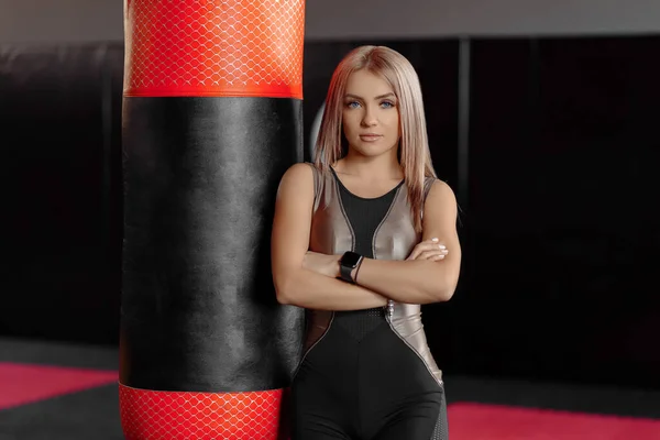 Selektivt fokus av blond kvinna i sportkläder när du står bakom boxningssäck. Ung kvinnlig fighter poserar i tomt gym, tittar på kameran. Begreppet boxning. — Stockfoto