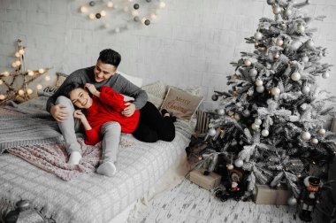 Kışlık kazak giyen neşeli çift rahat bir yatakta uzanıp gülüyor.