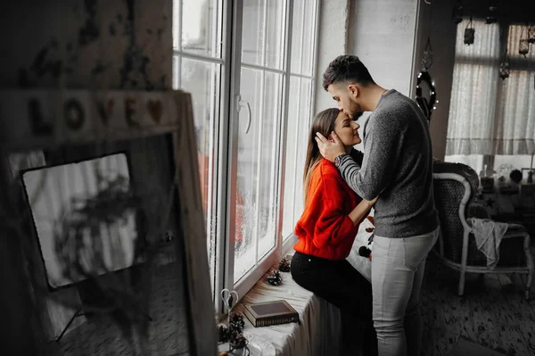 Young man in love kissing his girlfriend on the forehead while standing by the window