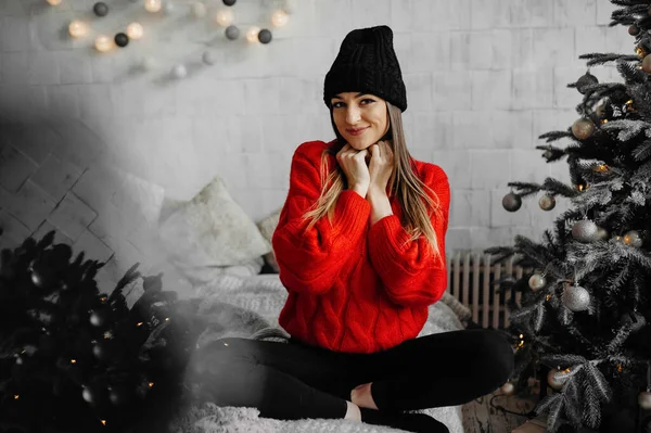 Yatakta bağdaş kurup Noel 'in tadını çıkaran, süveterli, mutlu genç bir kadının fotoğrafı.