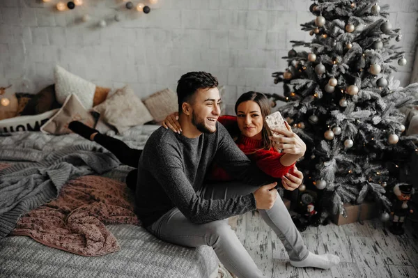 Mutlu genç çift evde selfie çekiyor, eğleniyor ve Noel ağacının yanında gülüyor.