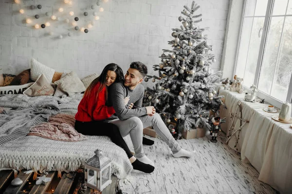 Happy couple in winter sweaters having fun lying on cozy bed and laughing, woman hugging her boyfriend spending winter holidays at home in warm christmas atmosphere — Stock Photo, Image