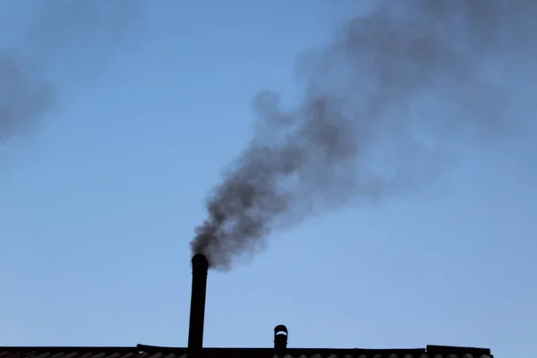 Bir köy evinin bacasından açık mavi gökyüzüne karşı siyah duman yükseliyor.