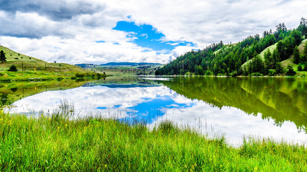 Blue Sky, Clouds and surrounding Mountains reflecting on the smooth water surface of Trapp Lake, located along Highway 5A between Kamloops and Merritt in British Columbia, Canada