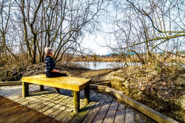 Kıdemli kadın, güzel bir kış gününde Fort Langley, British Columbia, Kanada yakınlarındaki Glen Valley Bölge Parkı 'nda Poplar Bar Iki-bit Bar iz yürüyüş ahşap bir tezgah üzerinde dinlenme