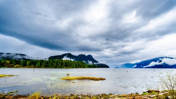 Oil slick on Pitt Lake under a dark cloudy sky with rain clouds hanging around the Mountains of the Coast Mountain Range in the Fraser Valley of British Columbia, Canada