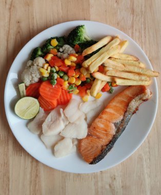 Izgara somon balığı ve çeşitli sebzelerle süslenmiş fırınlanmış morina balığı çeşitli sebzelerle bezelye limonlu patates kızartması ve ahşap masadaki beyaz tabak üzerinde soslar uluslararası taze market restoranı için yemek stilisti, menü reklam geçmişi.