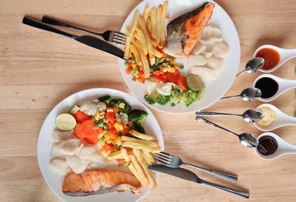 Grilled salmon fish and baked cod fish decorated with various vegetables corn carrot peas lemon French fries and sauces on white flat plate table on wooden table, food stylist for international fresh market restaurant, menu advertising backgrounds — Stock Image Grilled Salmon Fish Baked Cod Fish Decorated Various Vegetables Corn Stock Picture